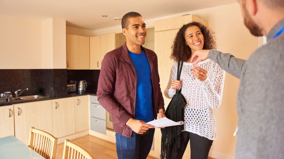 Two new, smiling renters stand in a kitchen while a landlord hands them keys.